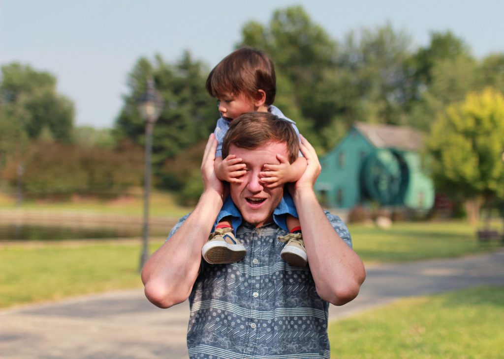 adult holding child on shoulders and covering adults eyes